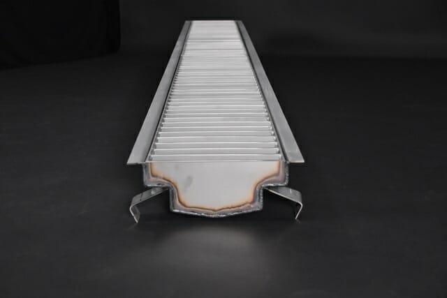Metallic grill with parallel slats, featuring a welded edge and support brackets, set against a dark background.