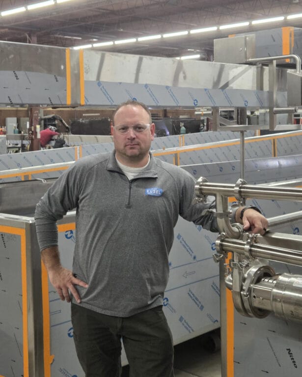 Man in a gray Kusel sweatshirt stands confidently beside industrial equipment in a workshop, wearing safety glasses.