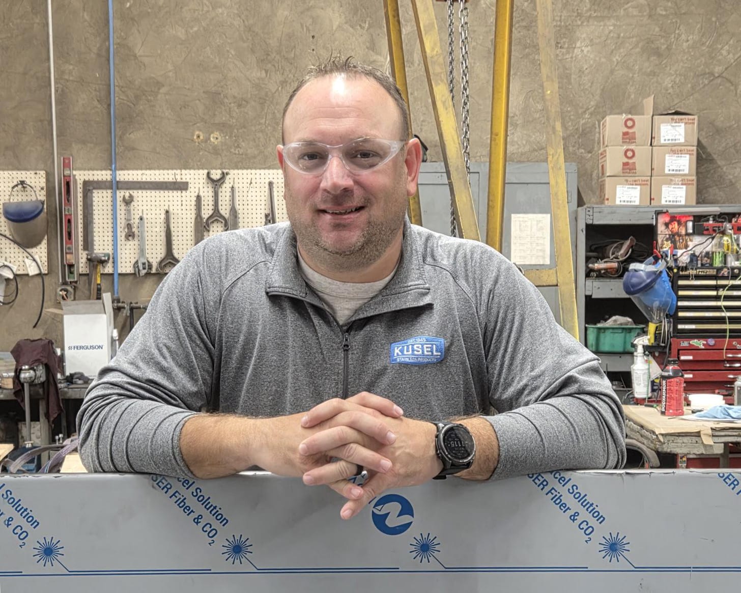 Man in safety glasses smiles while leaning on a workbench in a workshop filled with tools and equipment.