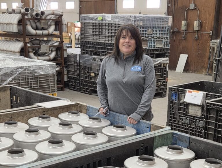 Woman in gray sweatshirt with "Kusel" logo stands in warehouse with metal parts.