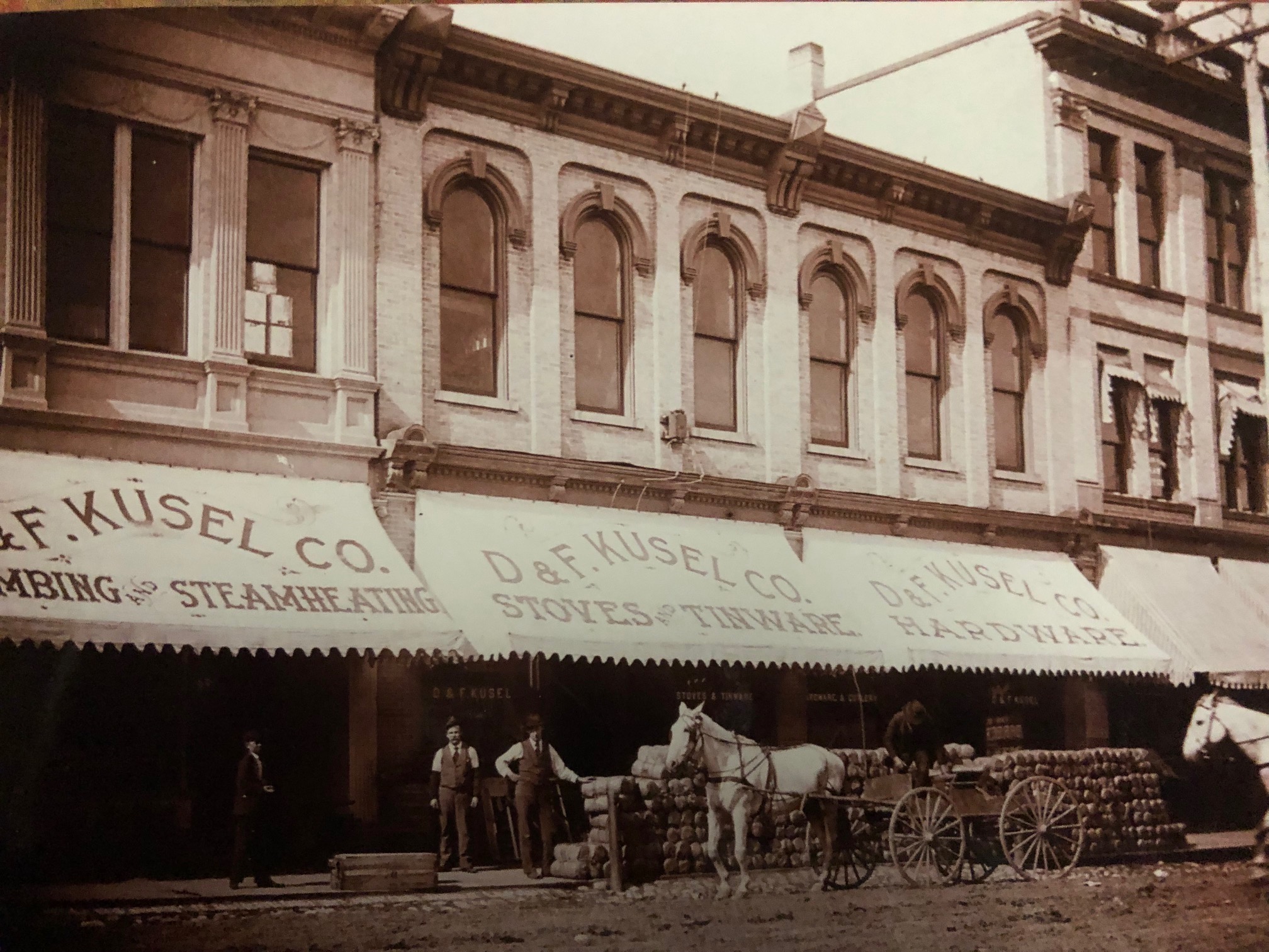 Vintage storefront with D&F Kusel Co. awnings, horse-drawn cart, and men.